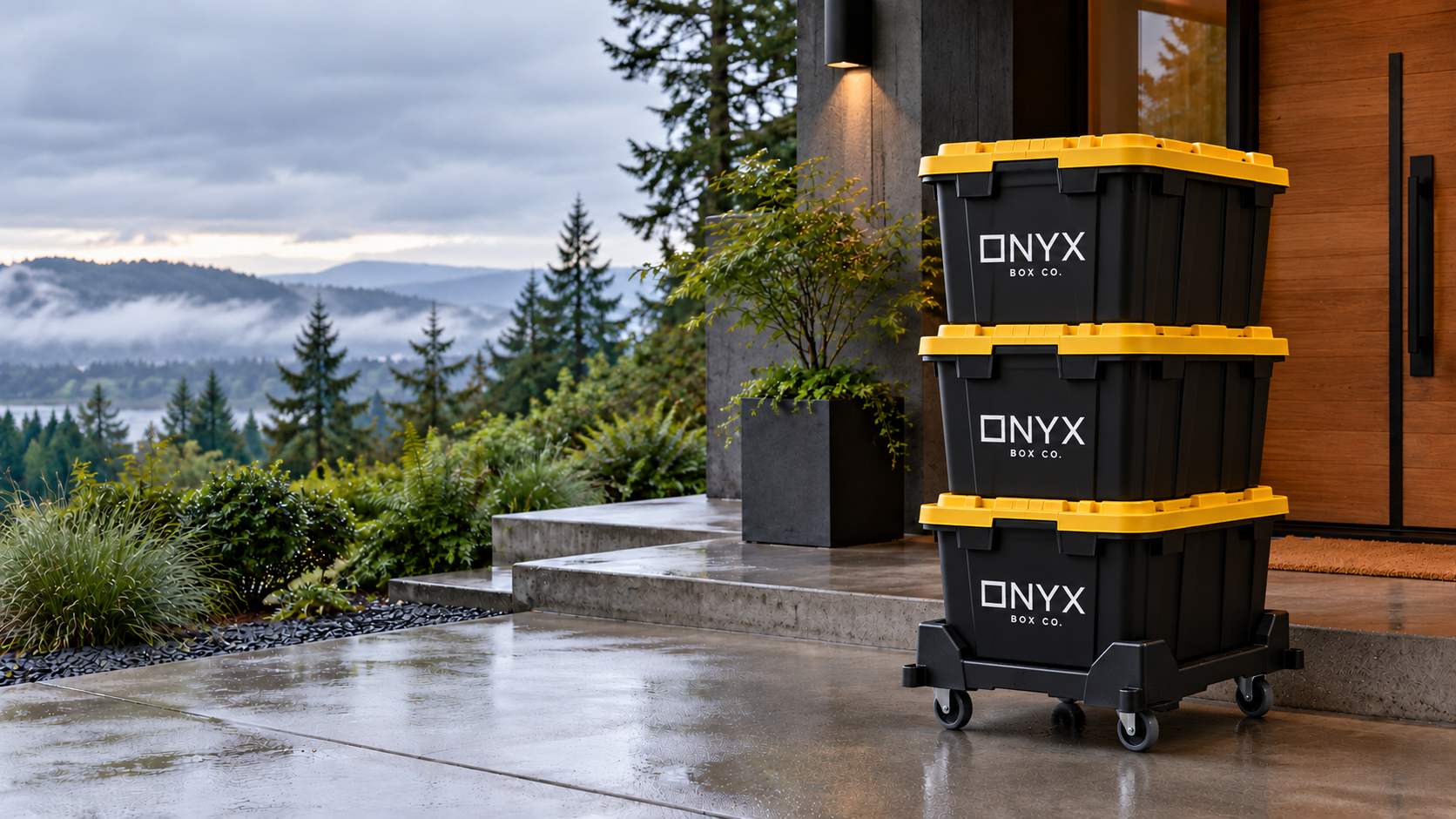 Three heavy-duty black Onyx Box Co. reusable moving totes shown narrow-end forward with gold lids, stacked on a fitted Tote Taxi outside a modern Pacific Northwest home